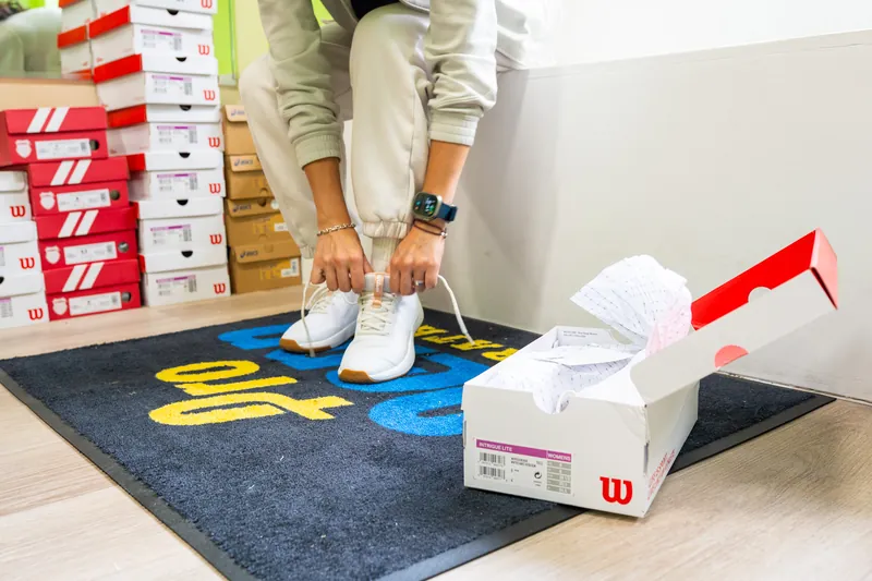 A customer lacing up a pair of Wilson tennis shoes in the shop, with K-Swiss, Wilson and Asics shoe boxes stacked behind