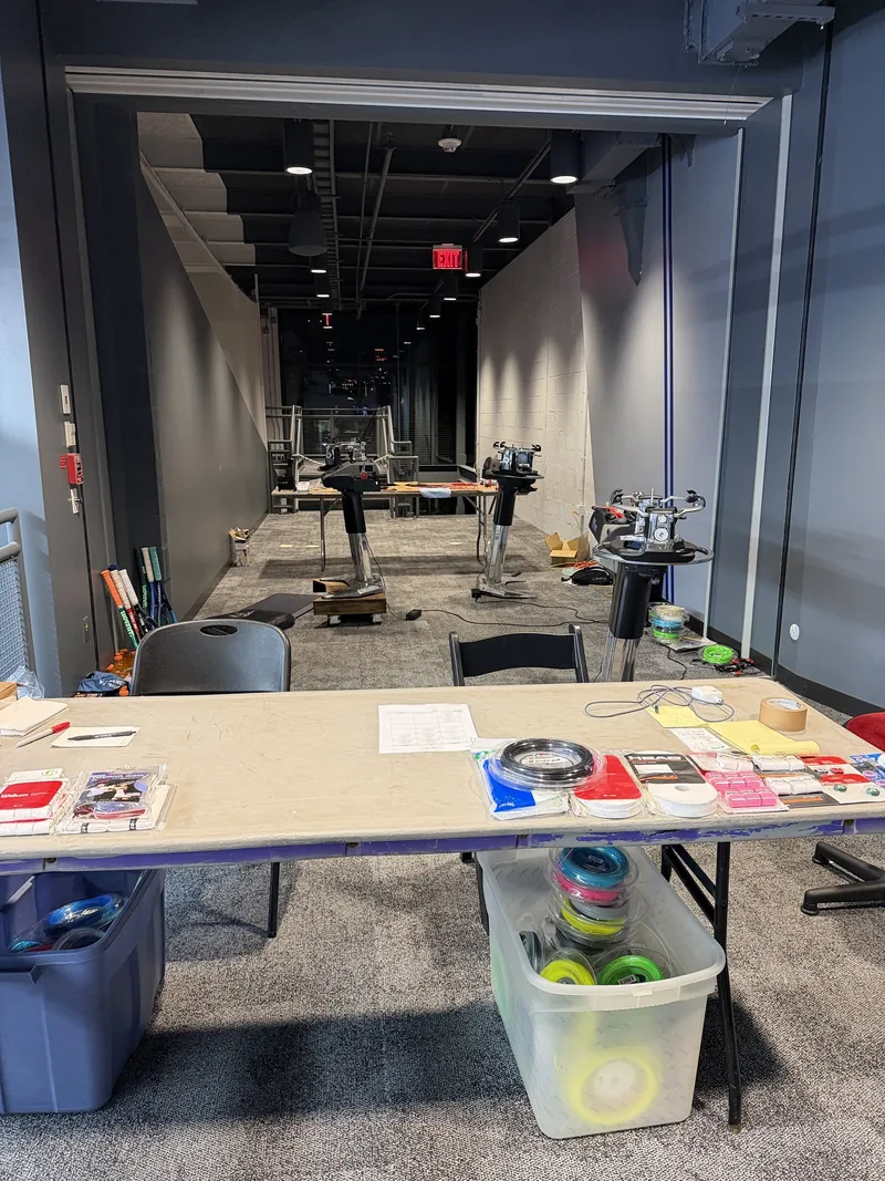 Inside a tournament stringing room: the drop-off table with string, grips and the log sheet in front, Wilson Baiardo stringing machines behind